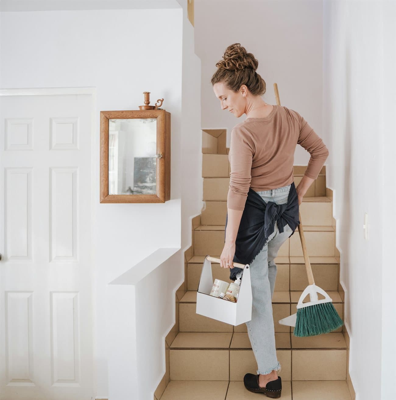 A woman carrying a cleaning caddy and broom up the stairs of a home